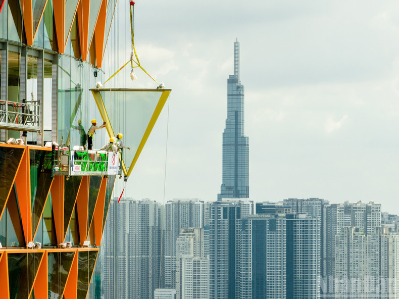 Photo: Construction at Great Heights by Khoi Dang (This image captures an impressive moment at a skyscraper construction site, where workers are suspended on a hanging scaffold, working in unison to install a massive glass panel onto the façade of a modern building distinguished by its bold glass design. In the distance stands a high-rise tower, an emblem of Ho Chi Minh City’s growth and dynamic rise.) anh4.jpg