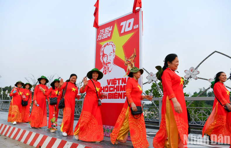 Women dressed in Ao Dai featuring the national flag of Vietnam passing by a poster celebrating the 70th anniversary of Dien Bien Phu Victory.