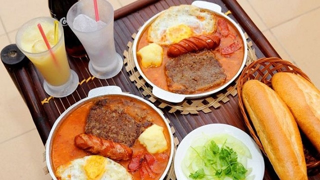 A combination of eggs, sunny side up, sausage, and pate is cooked in pan and served in the pan with a baguette.