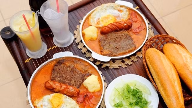 A combination of eggs, sunny side up, sausage, and pate is cooked in pan and served in the pan with a baguette.