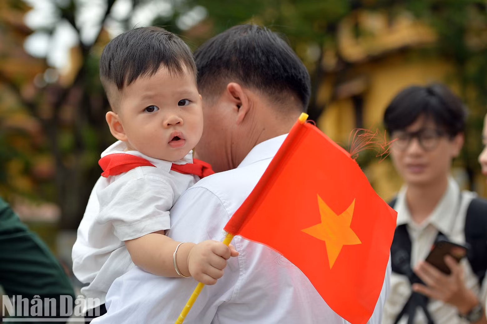 A young boy wearing a red scarf holds a red flag in his hand.