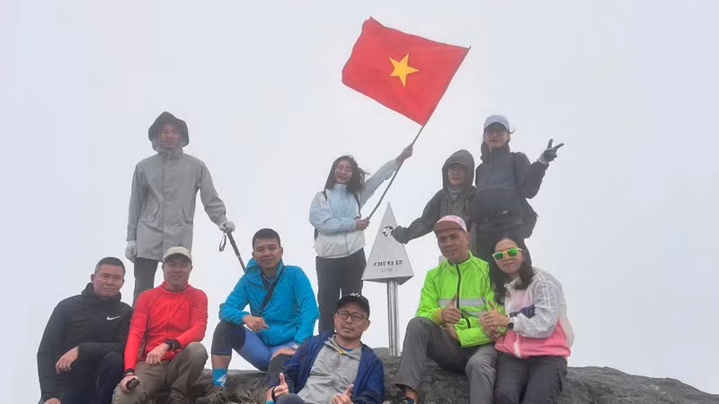 A group of tourists pose with the metal pyramid marked “Chu Va 2,751m” amidst howling winds.
